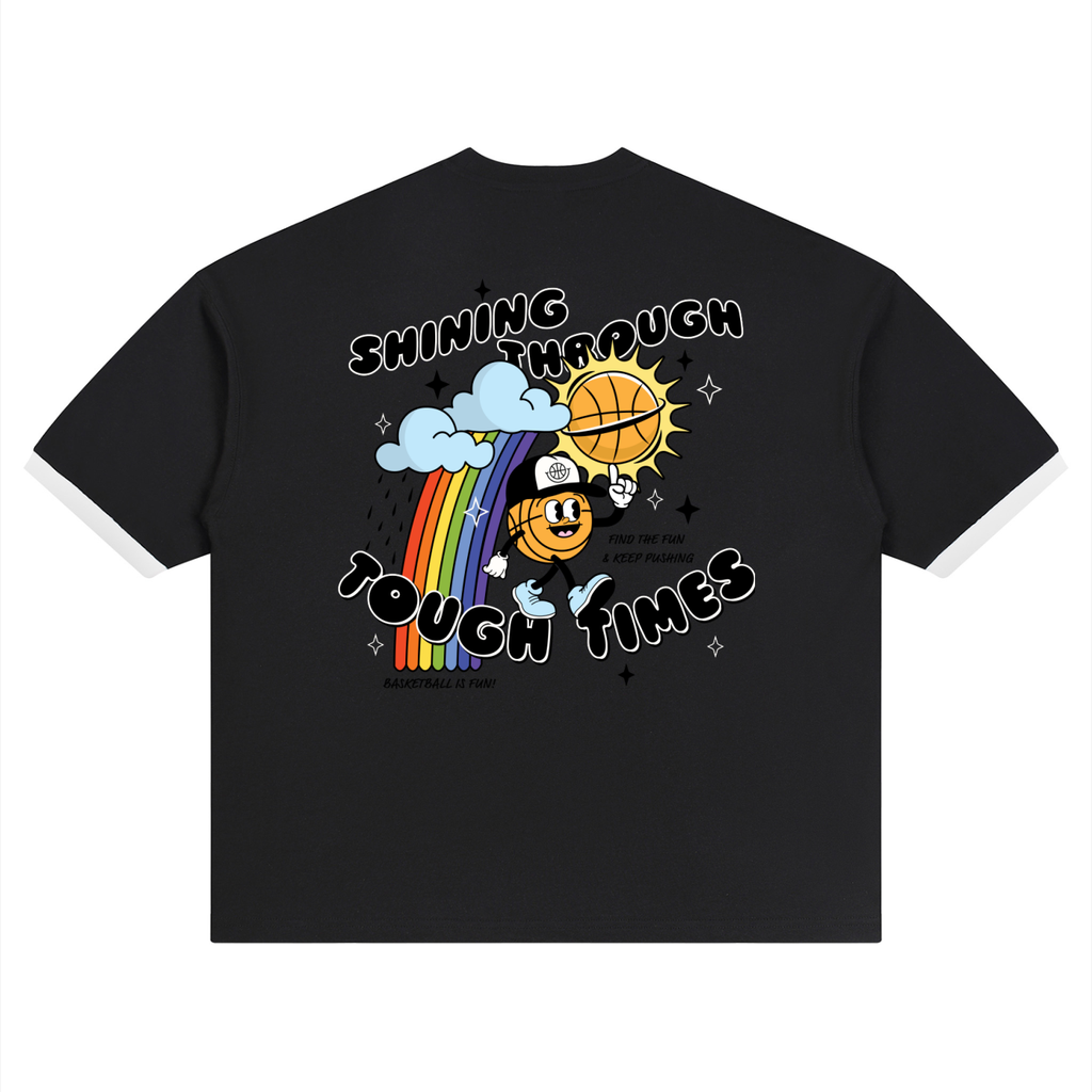 A black short-sleeve t-shirt with white contrast stitching on the sleeves and a colorful graphic on the front. The graphic features a cartoon basketball mascot holding a basketball under a rainbow and clouds, with the text 'SHINING THROUGH TOUGH TIMES'.