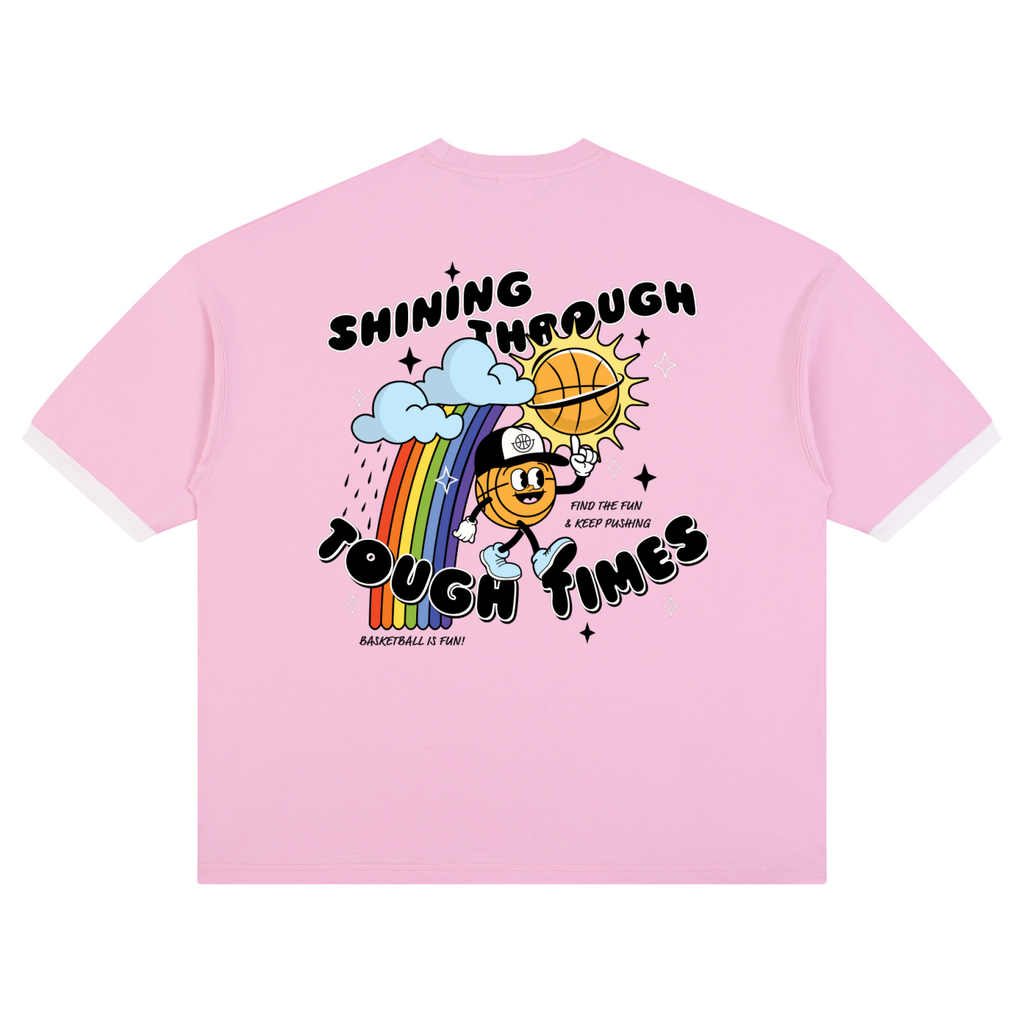 A light pink short-sleeve t-shirt with white ribbed trim on the collar and cuffs, featuring a colorful graphic on the back with a cartoon character, rainbow, basketball, and the text 'SHINING THROUGH TOUGH TIMES'.