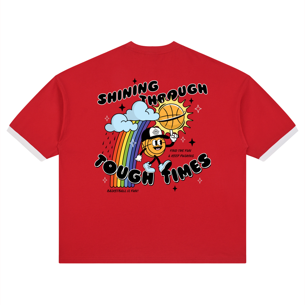 A red short-sleeve t-shirt with white trim on the sleeves and collar, featuring a colorful graphic on the front with a cartoon basketball character, a rainbow, clouds, and the text 'SHINING THROUGH TOUGH TIMES'.